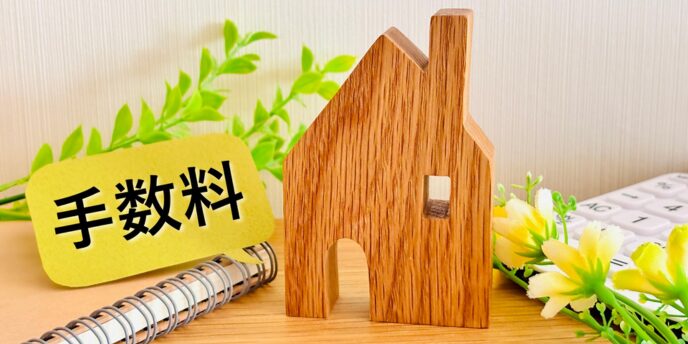 「手数料」の札と木製の家の模型、花や電卓が並ぶ画像