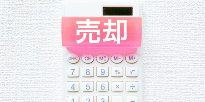 テクスチャー壁に掛けられた白い電卓の上部に、ピンクの付箋で「売却」と書かれているシンプルなビジネスイメージ