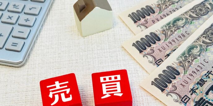 電卓と小さな家の模型、千円札が並ぶテーブル上に「売」「買」と書かれた赤いブロックが配置された不動産取引イメージ