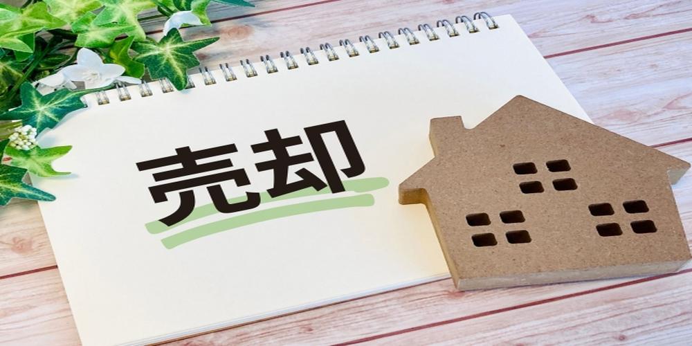 売却と書かれたノートと家の模型が並ぶ木目調の背景