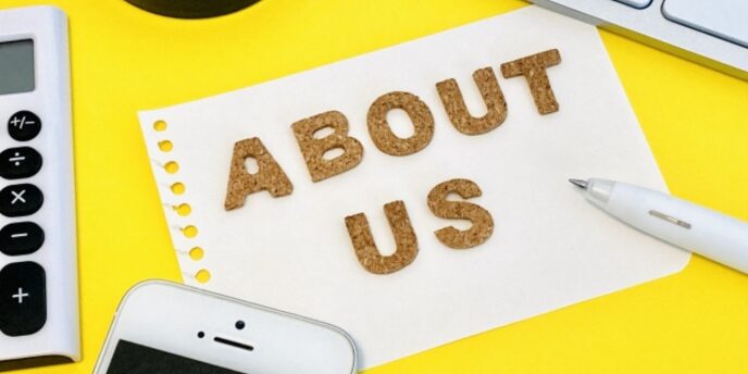 黄色い背景の上に置かれた白いメモ用紙に「ABOUT US」と書かれ、周囲に電卓やスマートフォンがある画像