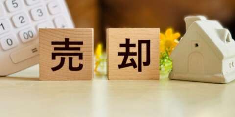 木製ブロックに「売却」と書かれ、横に家のミニチュアと電卓、花が置かれている画像