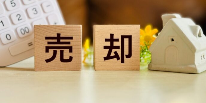 木製ブロックに「売却」と書かれ、横に家のミニチュアと電卓、花が置かれている画像
