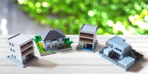 木目調のテーブルに4種類の住宅模型が並べられている画像。背景は緑のボケが広がる自然の景色