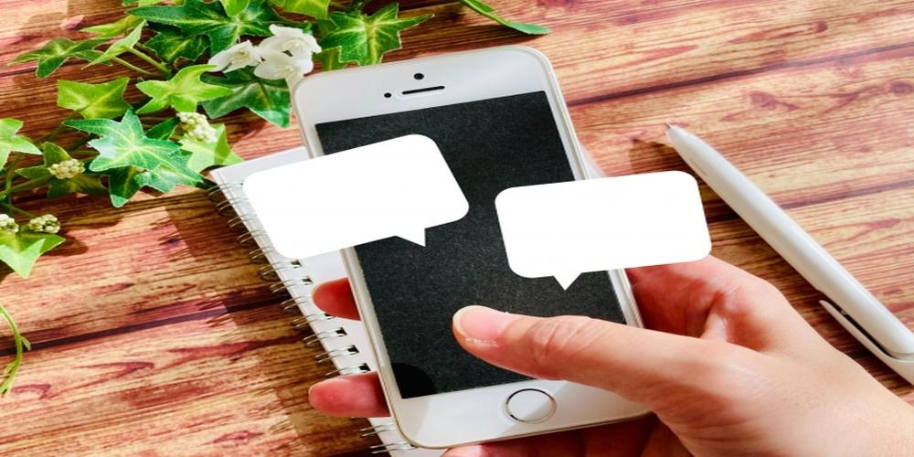 吹き出しが表示されたスマートフォンを持つ手と木目調のデスク上のペンと植物